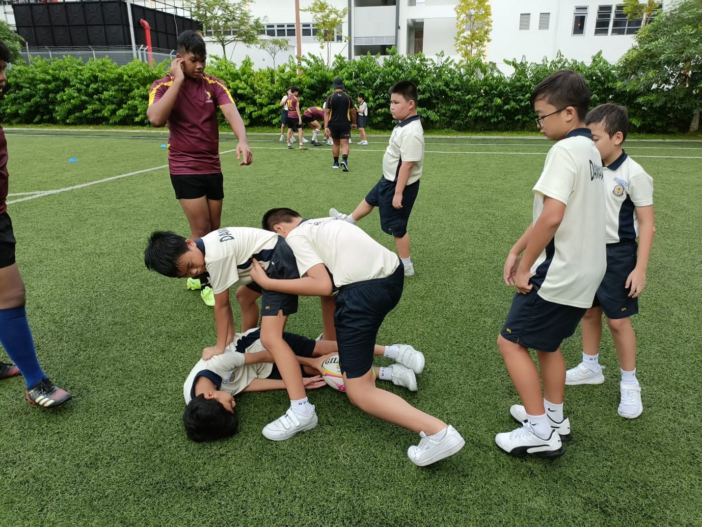 Young boys in school uniforms playing rugby on a green field, with one player tackled to the ground holding the ball.
